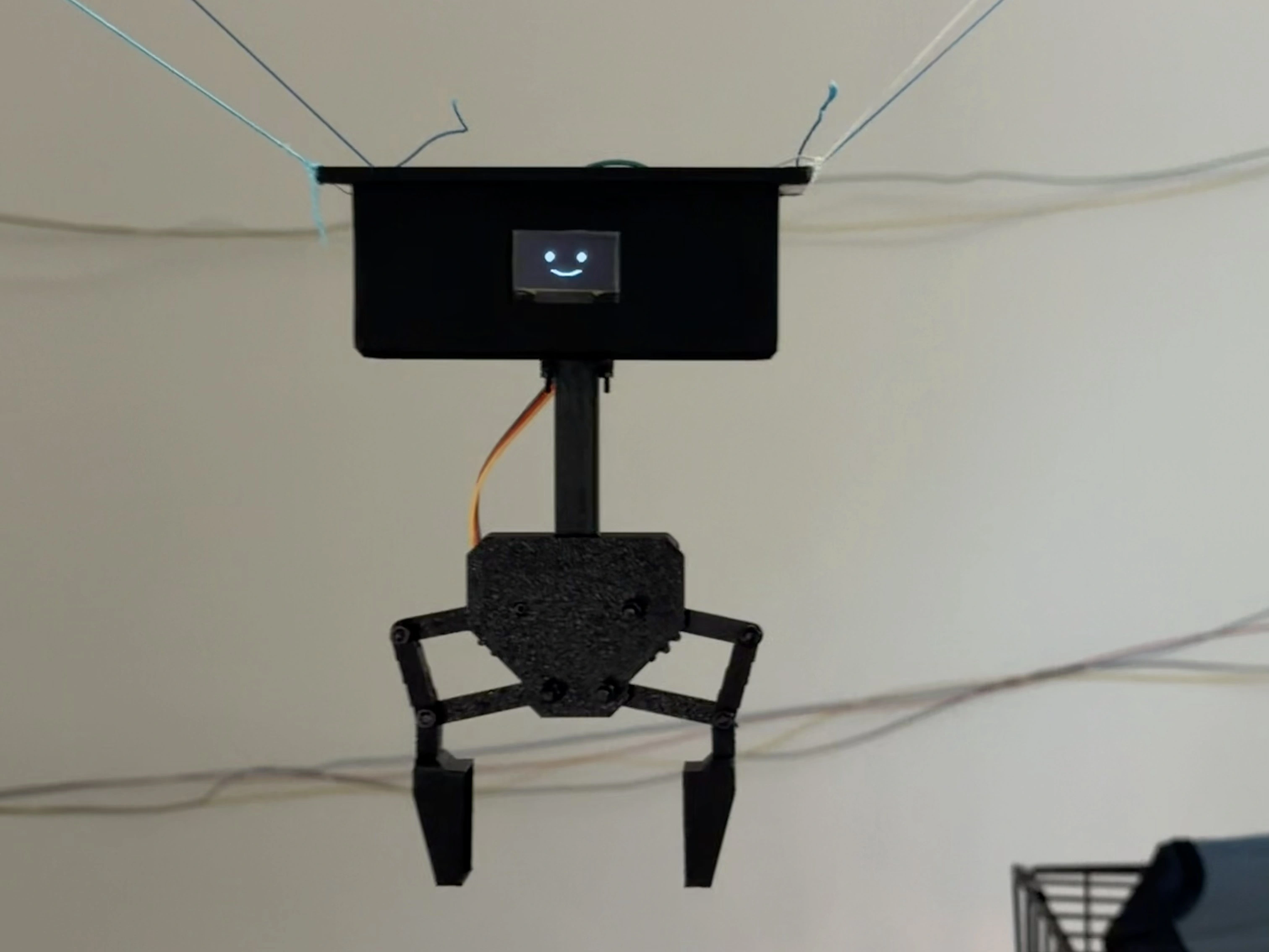 The platform hanging from its four lines: a black printed box with a small OLED showing a smiling face, and a two-jaw claw on a stalk beneath it.